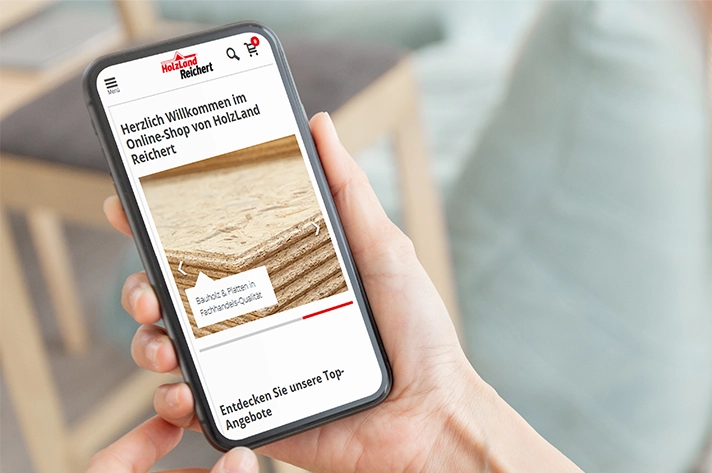 Smartphone zeigt die Startseite des HolzLand Reichert Onlineshops mit Holzplatten-Motiv.