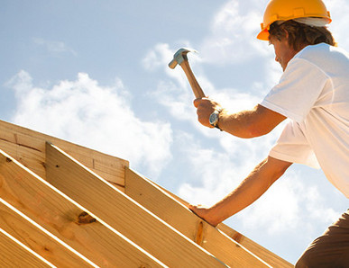 Dachdecker mit gelbem Schutzhelm beim Befestigen von Konstruktionsvollholz (KVH) auf einem Dachstuhl unter strahlendem Himmel.