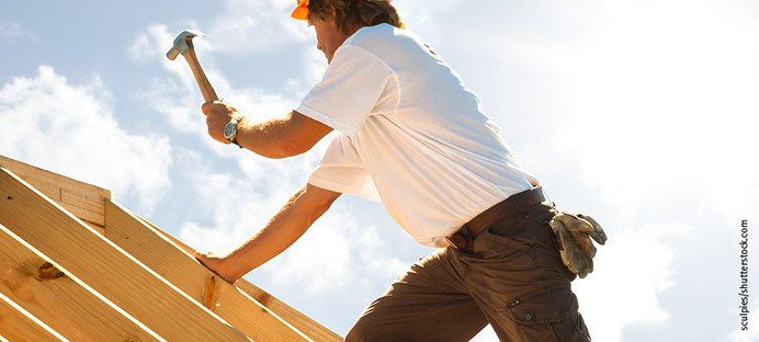 Dachdecker mit gelbem Schutzhelm beim Befestigen von Konstruktionsvollholz (KVH) auf einem Dachstuhl unter strahlendem Himmel.
