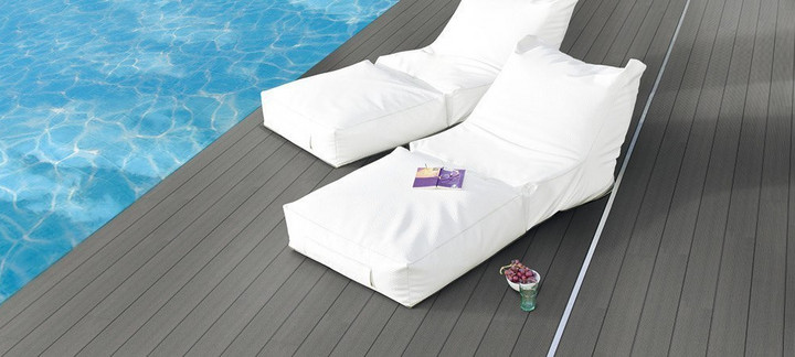 Elegante Poolterrasse mit dunklem WPC-Belag, zwei weißen Lounge-Liegen und ruhiger Wasserreflexion.